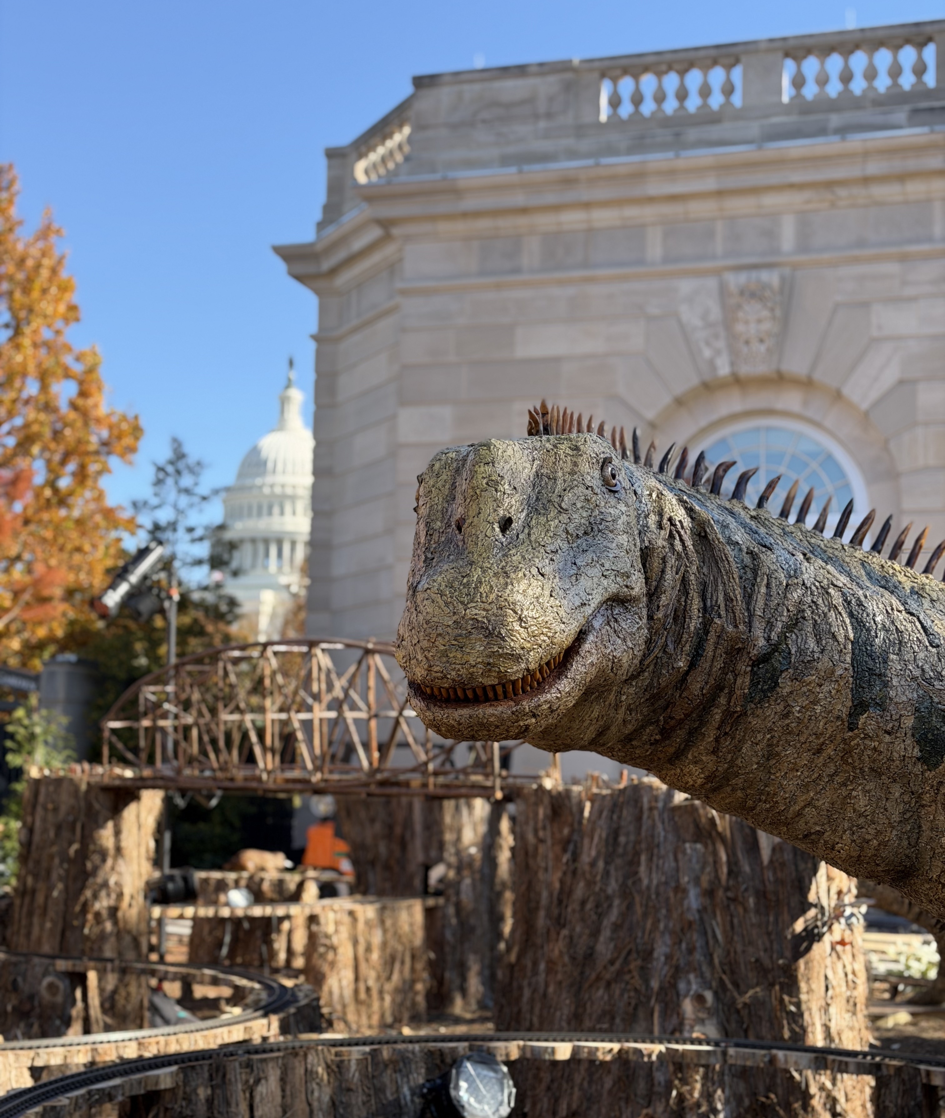 A model of a dinosaur made from plant parts in an ourdoor garden setting with model train tracks beneath and behind