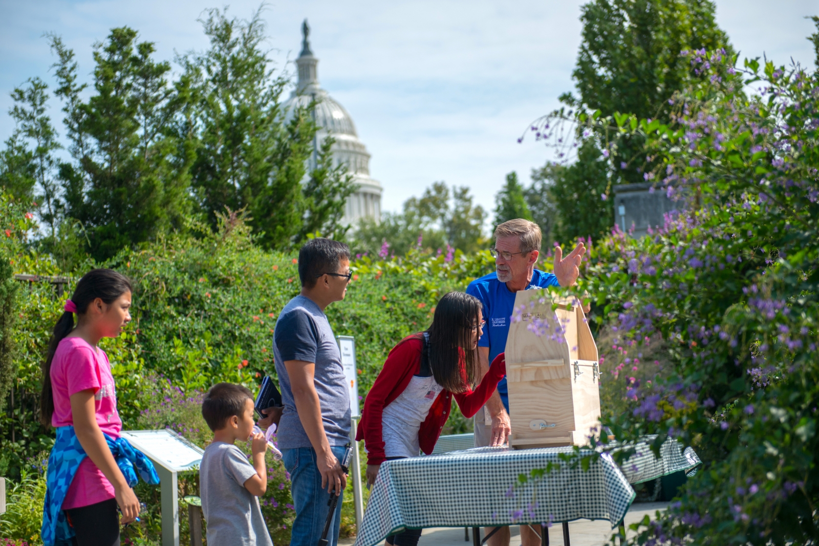 A teenage girl looks closely at a closed beehive, while family looks on. Flowering plants surround them