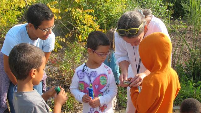 three young kids view a hand lens held by an instructor in an outdoor garden