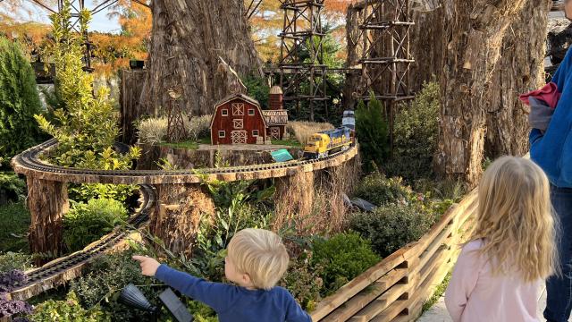 A young child points at a passing train in an outdoor display of model trails and plants