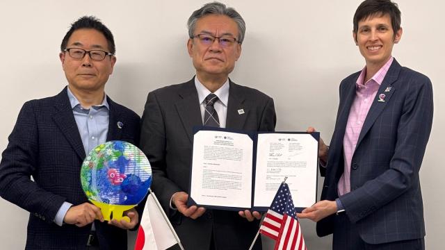 Three people in suits stand holding a signed agreement