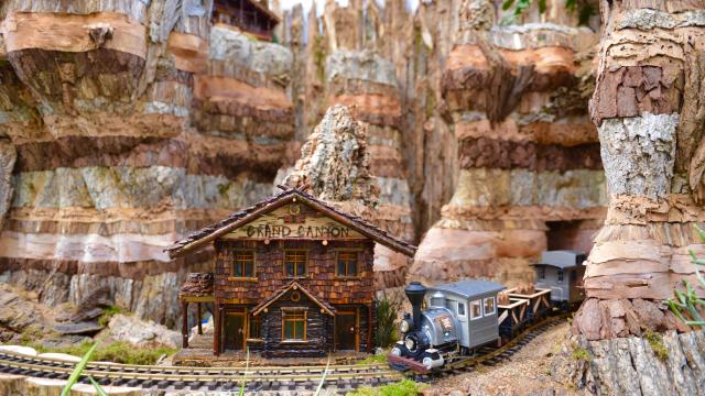 A model train with a silver engine and the National Park Service emblem runs along a track beside a model of the Grand Canyon Depot made from many different plant parts amidst a recreation of the Grand Canyon also made from many different plant parts