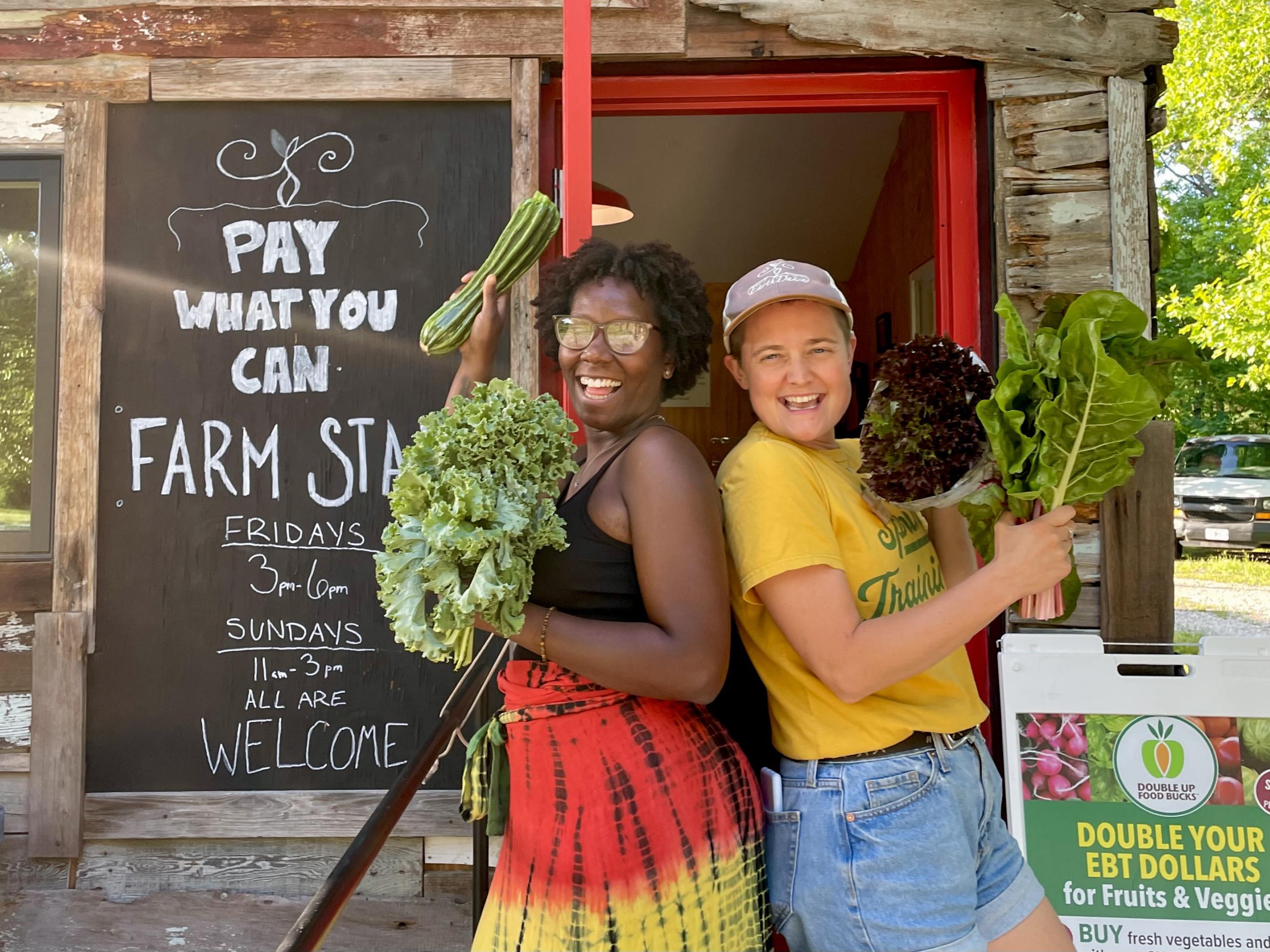 Two women hold green produce in front of a sign that says Pay What You Can Farm Stand