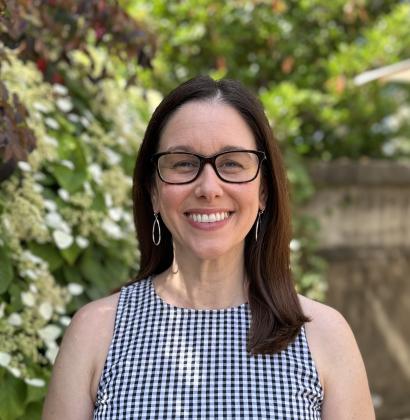 A woman with long brown hair and black glasses stands in an outdoor garden