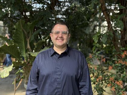 a man wearing glasses stands in a greenhouse room filled with green tropical plants
